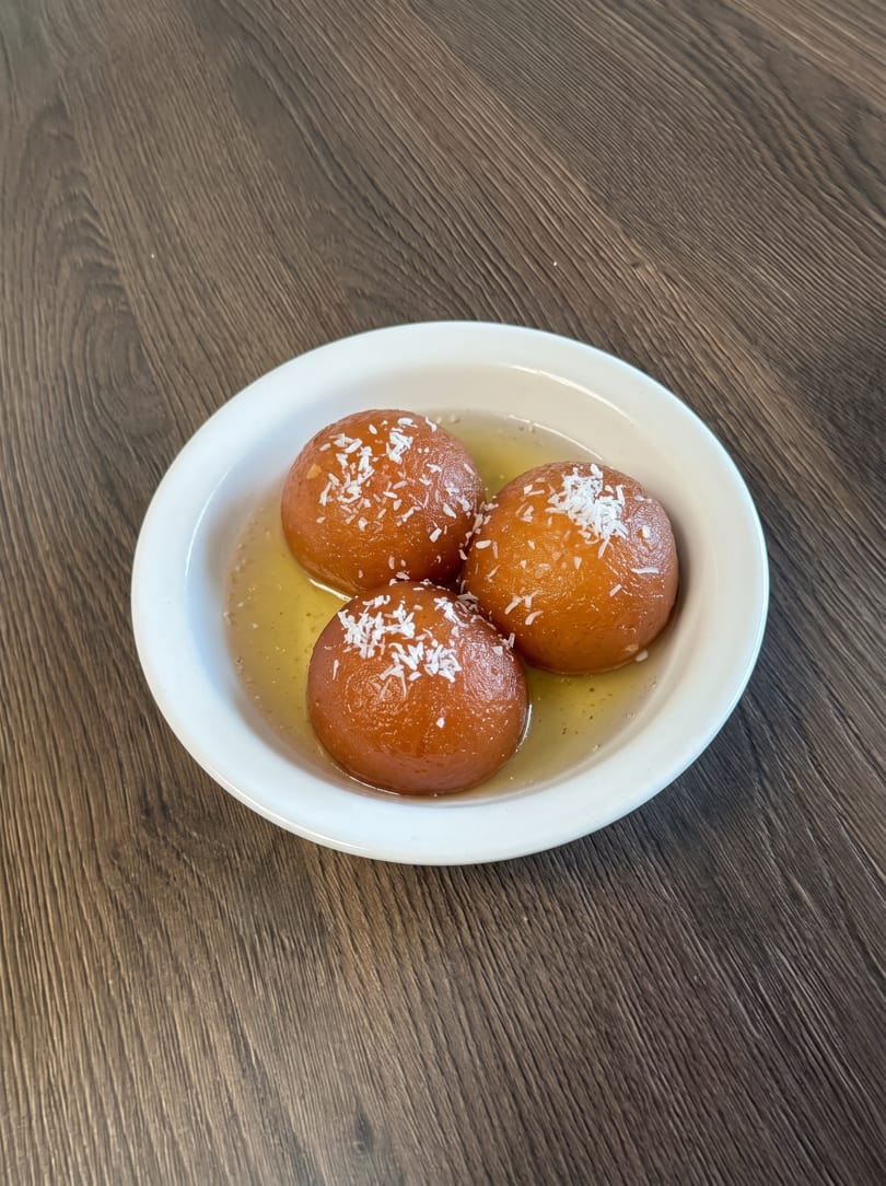 Three gulab jamun, Indian fried dough balls in syrup, topped with coconut flakes on a white plate.
