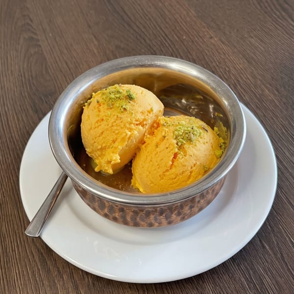 Two scoops of mango kulfi with pistachio sprinkles in a metal bowl on a white plate.