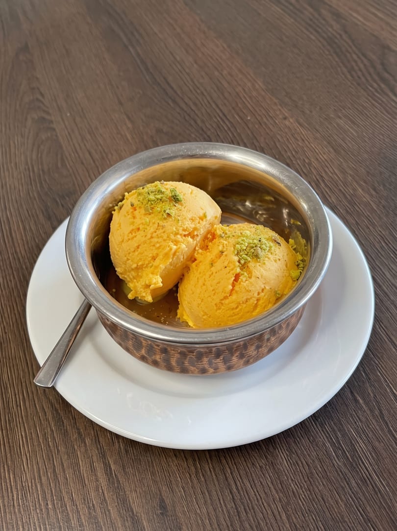 Two scoops of mango kulfi with pistachio sprinkles in a metal bowl on a white plate.