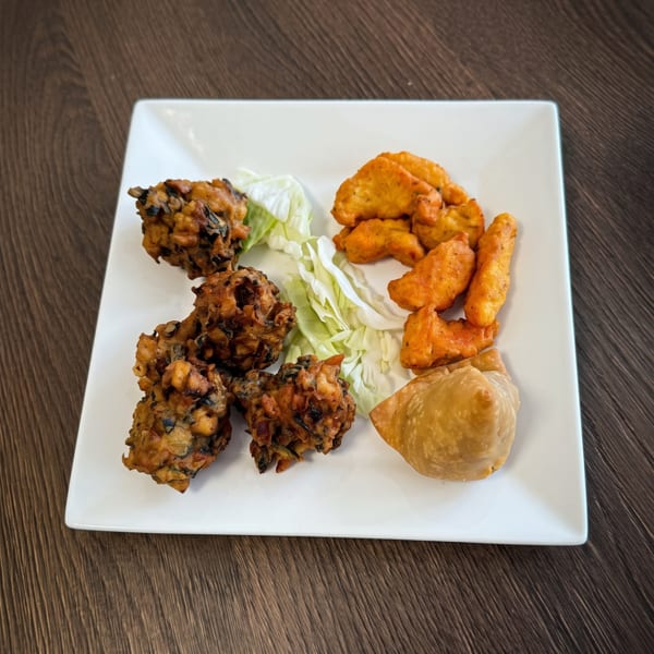 A white square plate with pakoras, chicken pakora, a samosa, and shredded lettuce on a wooden table.