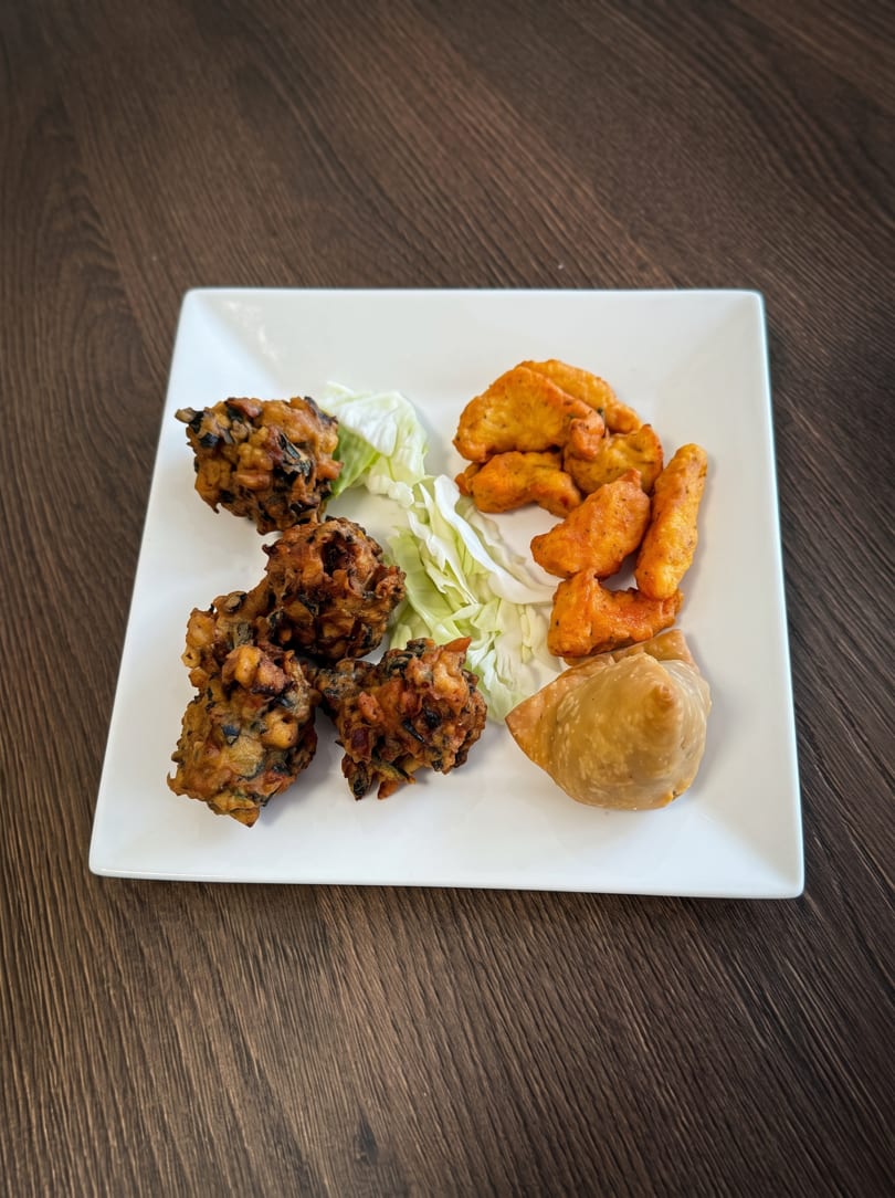 A white square plate with pakoras, chicken pakora, a samosa, and shredded lettuce on a wooden table.